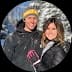 Smiling man in yellow beanie and woman in pink gloves pose against snowy trees.