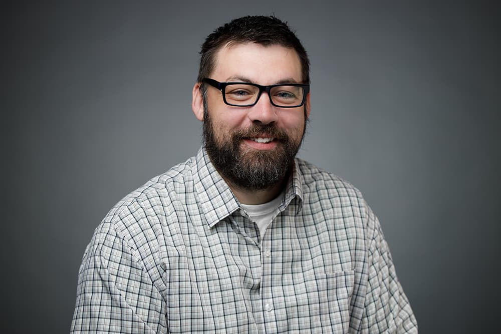 Smiling man with a beard and glasses wearing a plaid shirt against a grey background.