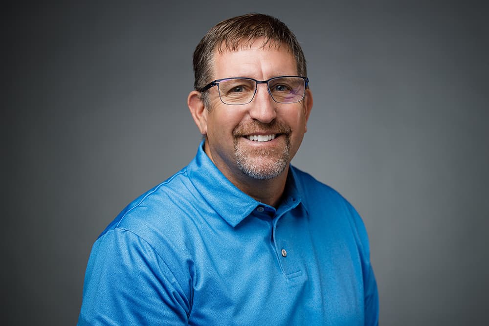 Smiling man with glasses and a goatee wearing a blue polo shirt against gray background.