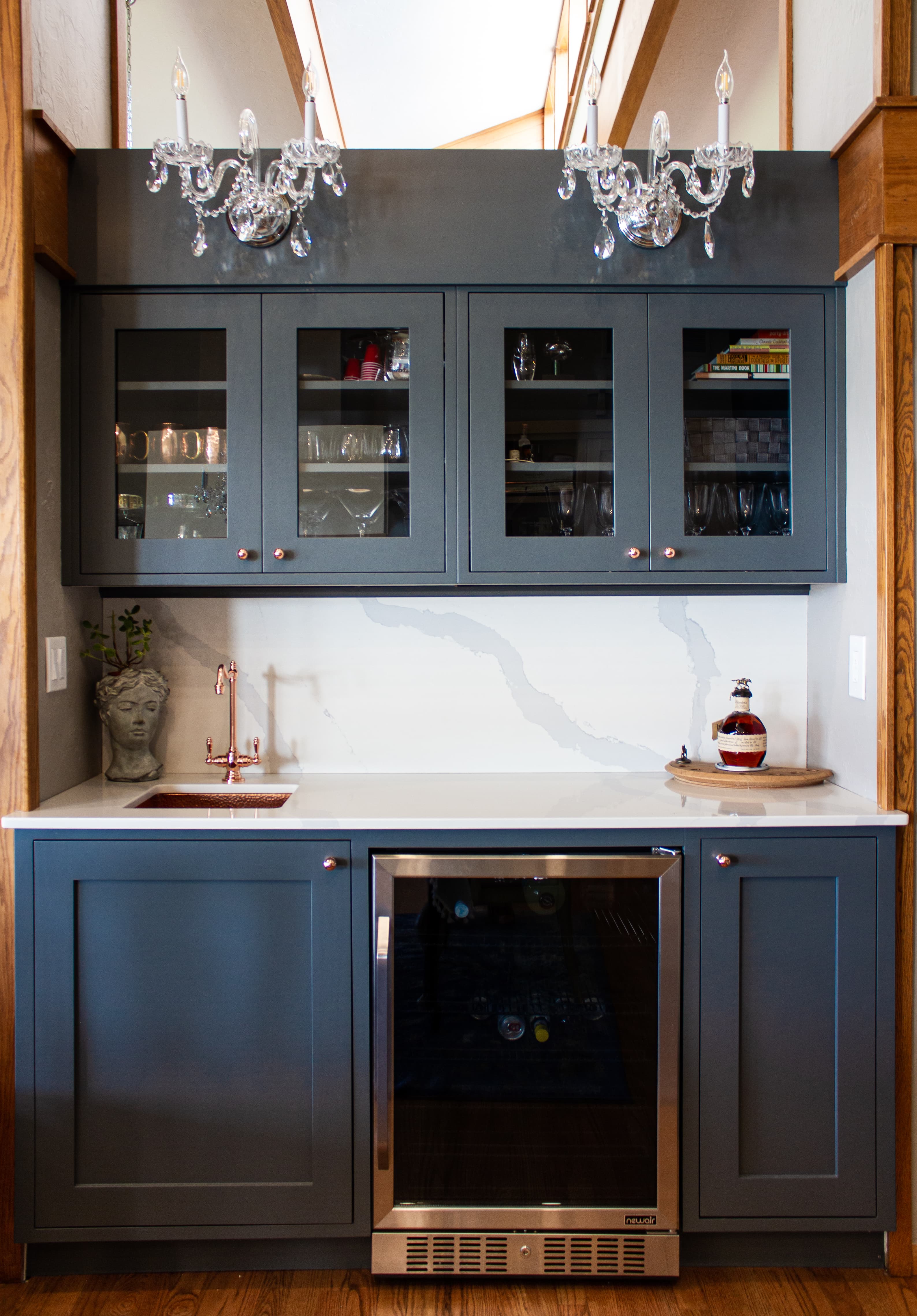 Modern home bar featuring grey cabinets, marble backsplash, wine fridge, copper sink, and crystal chandeliers.