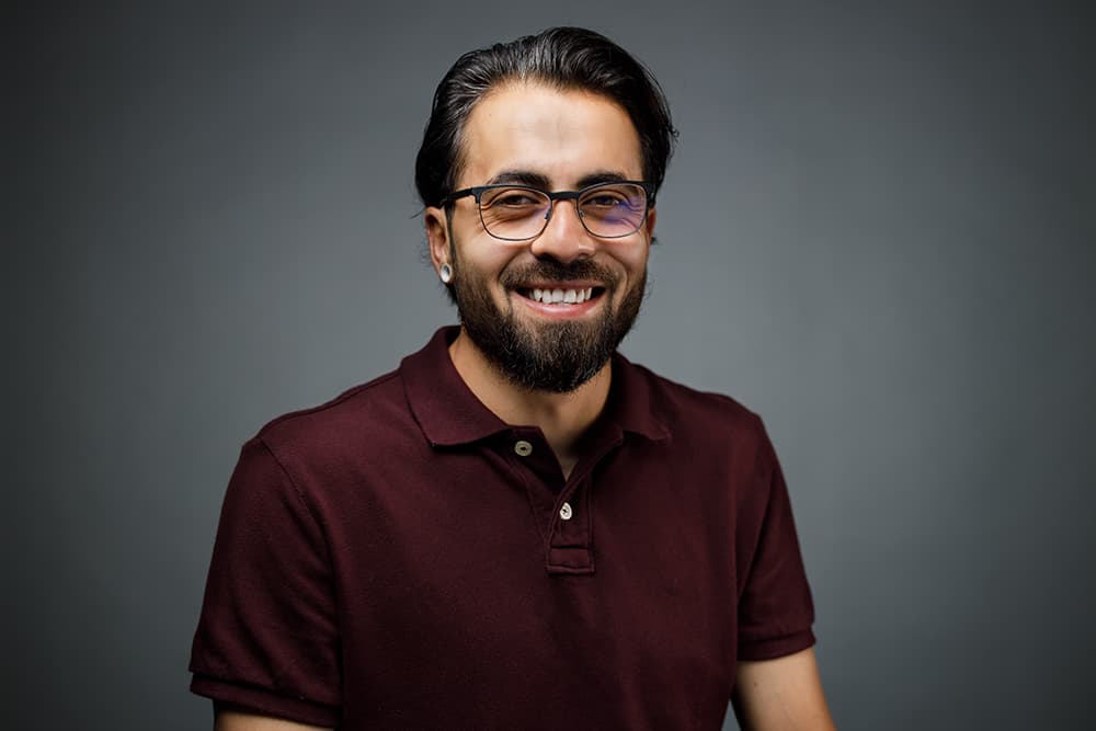 Smiling bearded man wearing glasses and a maroon polo shirt against a gray background.