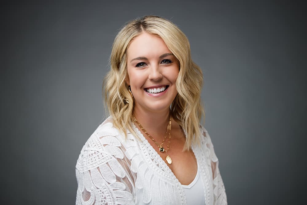 Smiling blonde woman with wavy hair wearing a white lace top and gold jewelry.
