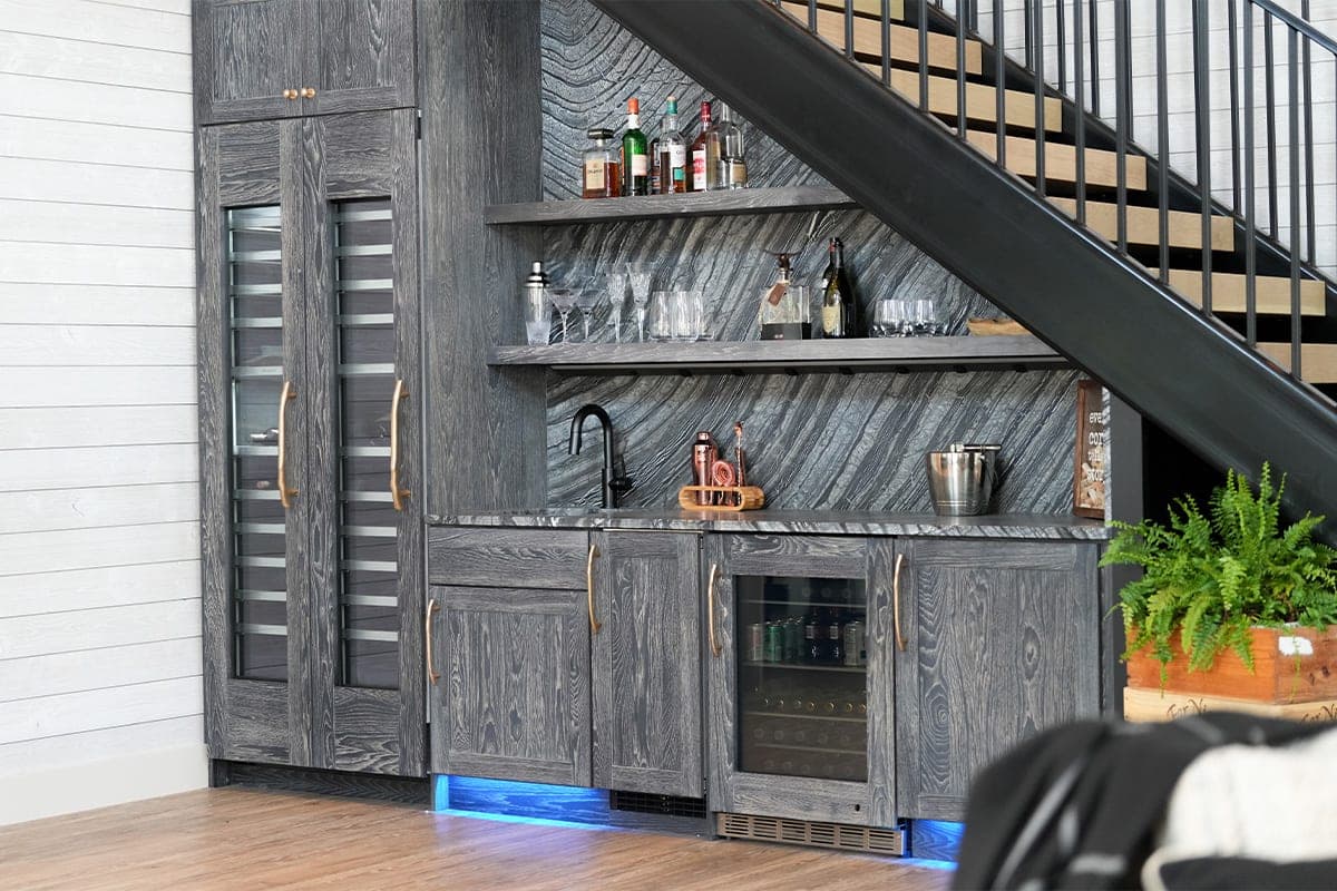 Modern home bar under stairs with dark grey cabinets, wine fridge, and blue LED lighting.