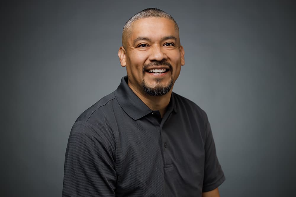 Smiling man with short hair and goatee wearing a gray polo shirt against gray background.