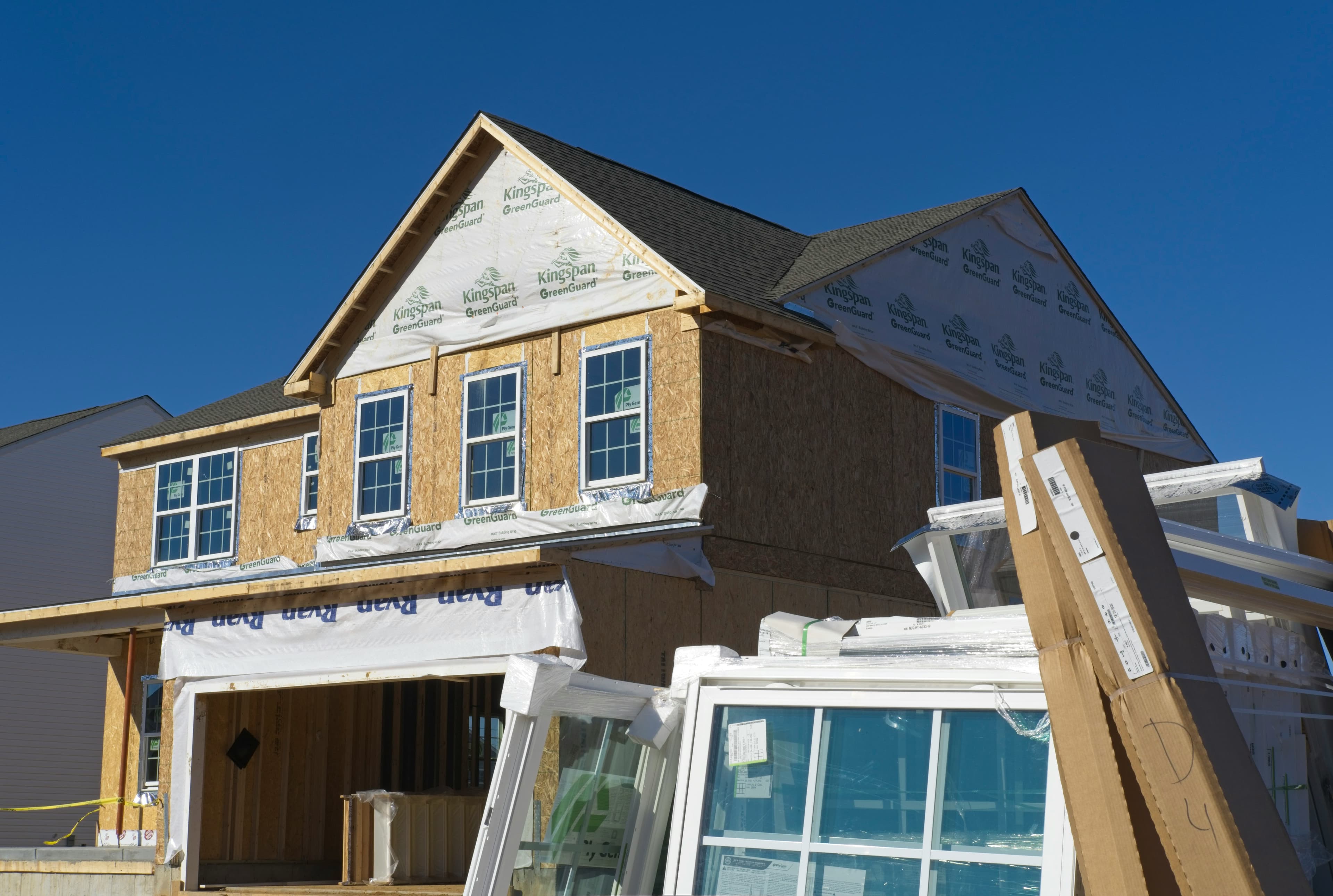 Two-story house under construction with house wrap and stacks of new windows in the foreground.