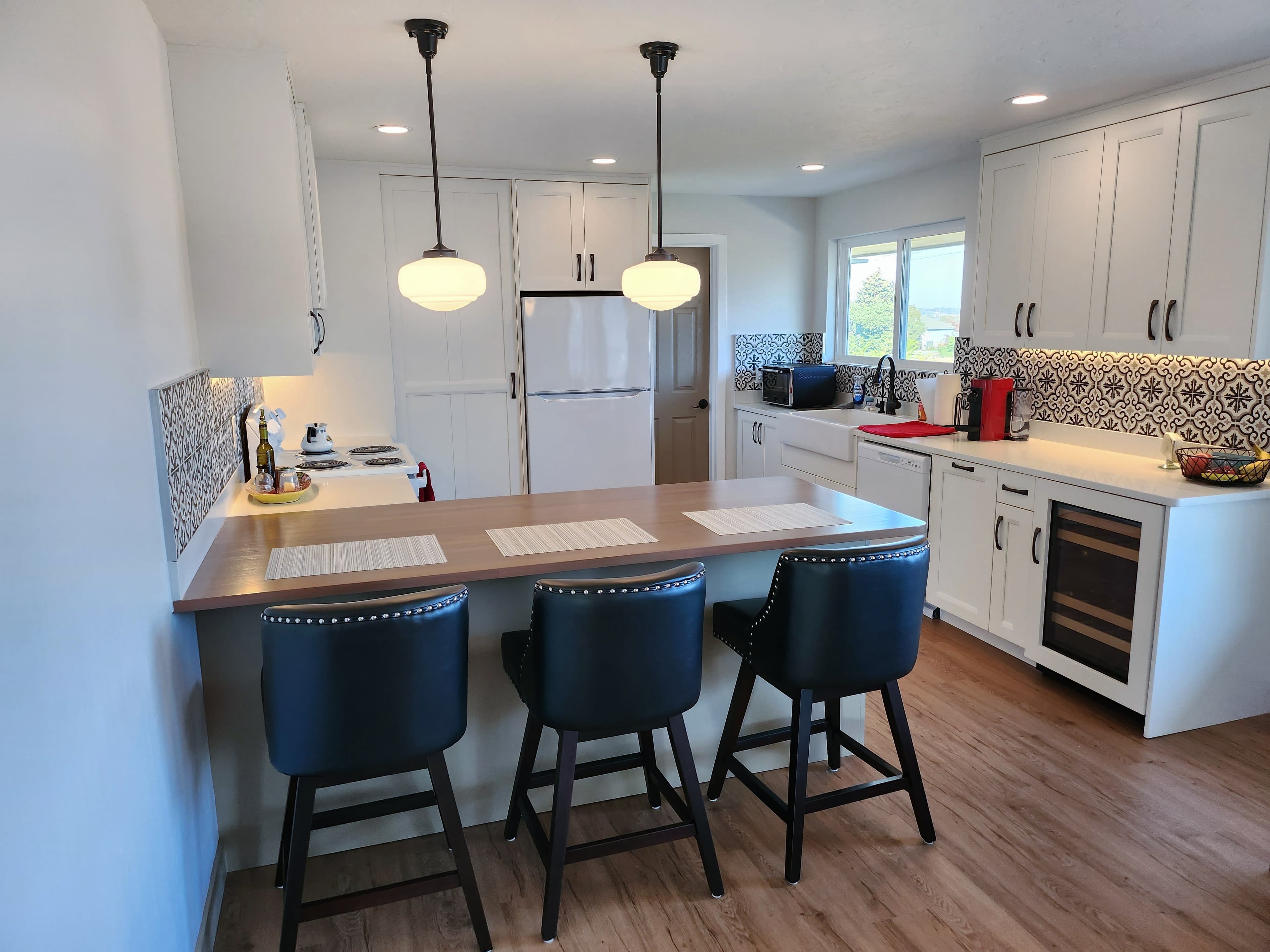 Modern white kitchen with wood island, three black leather stools, and patterned tile backsplash.