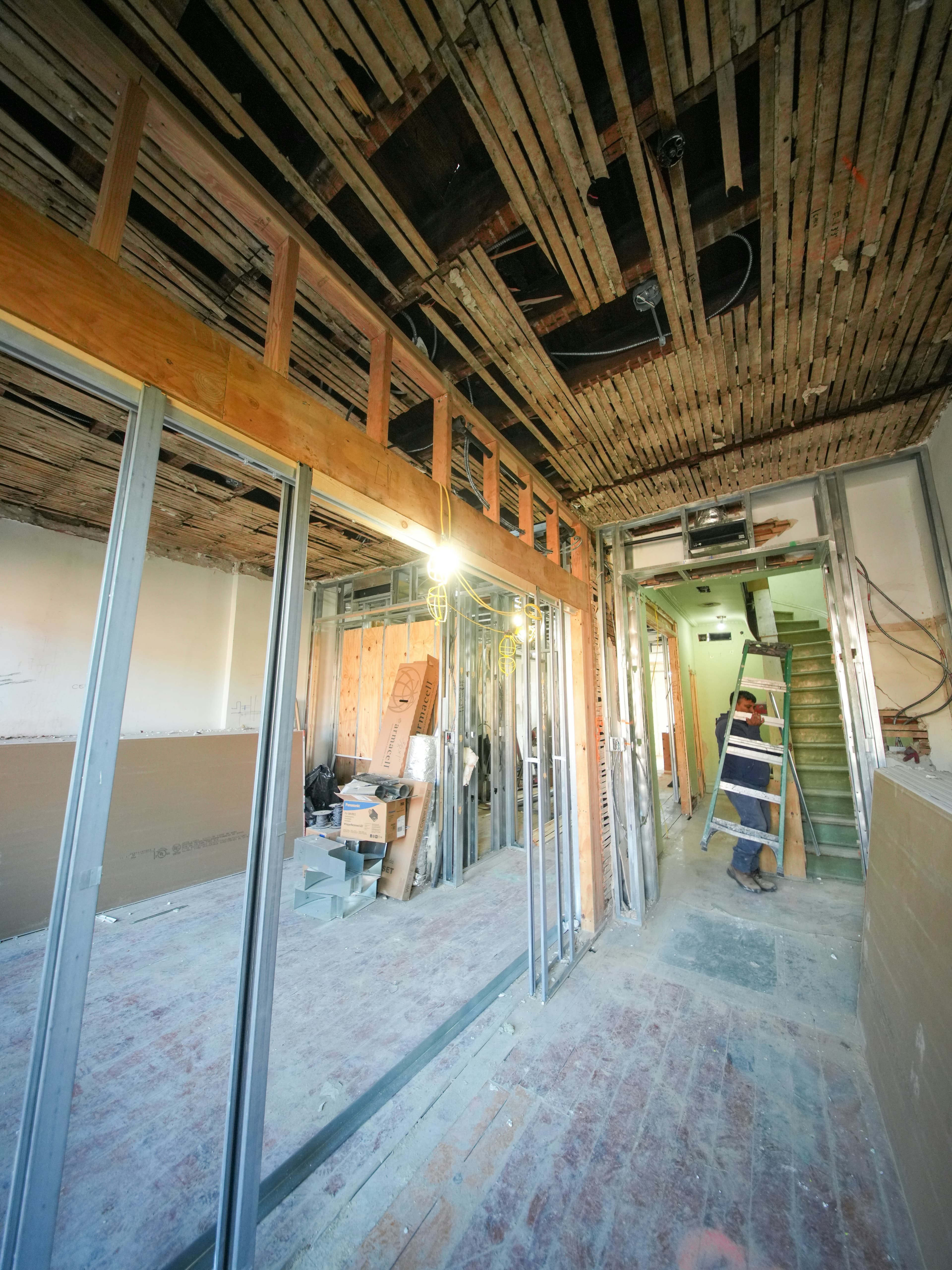 Interior renovation with exposed ceiling lath, metal framing, and a worker on a ladder.