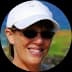 Smiling woman wearing a white baseball cap and dark sunglasses with a blurred coastal background.